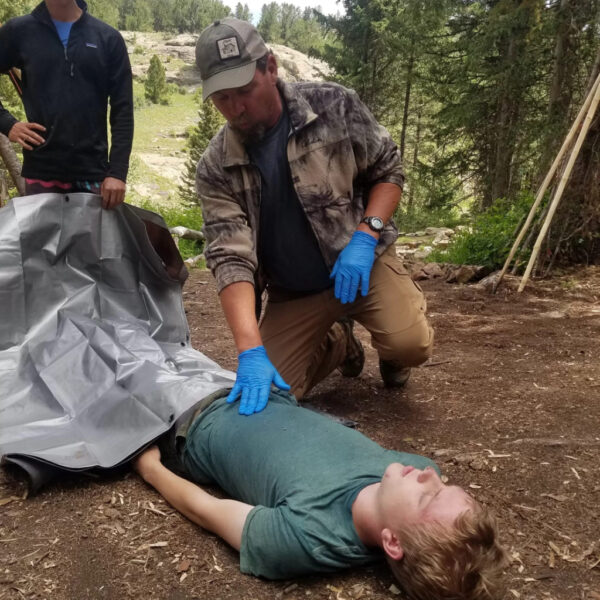 An outdoor wilderness first aid instructor wearing blue gloves checks the abdomen of a mock patient lying on the ground during a training scenario. Another student stands nearby holding a tarp, ready to assist with the simulated evacuation.