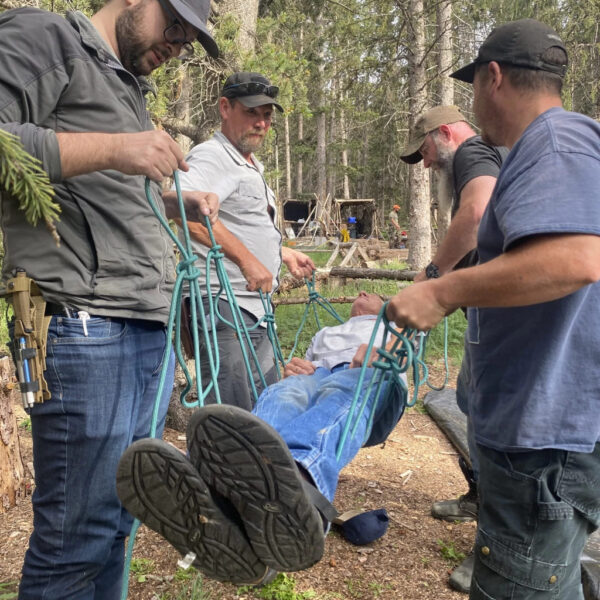 Four men practice a wilderness rescue scenario by lifting a person using rope techniques in a forested area. The person being carried appears calm, and the group is focused on coordinating the lift.