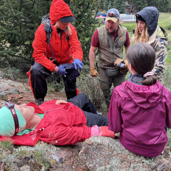 A group of wilderness first aid students in rain gear attend to a simulated patient lying on a rock. One student kneels to assess the patient, while others observe and assist during the outdoor scenario training in a forested campsite.