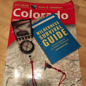 Wilderness Survival Guide book by Jason Marsteiner placed on a Colorado Atlas map alongside a baseplate compass with a red lanyard