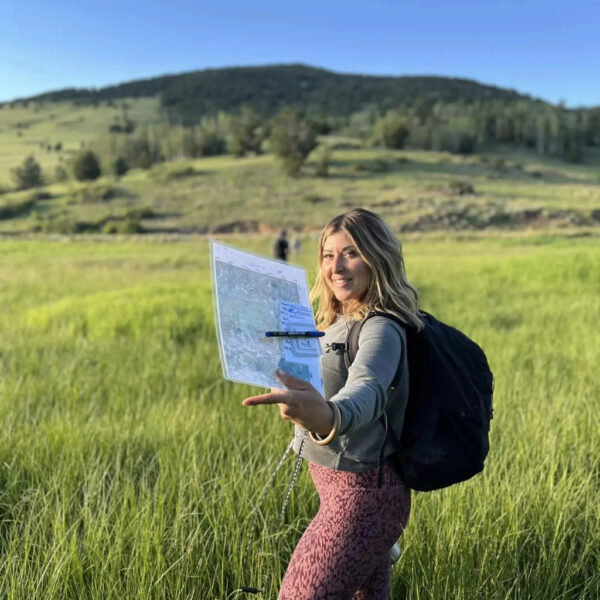 Smiling woman in hiking gear holds up a map while standing in a grassy field with hills behind her.