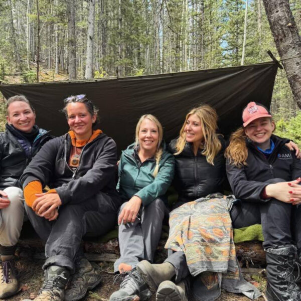 Group of five women smiling and sitting together under a tarp shelter in a forest during a Wildkind women’s outdoor retreat, wearing outdoor gear and surrounded by trees.