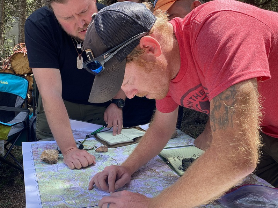 Matt Boger, The Survival University’s land navigation instructor, leans over a topographic map while teaching a land nav exercise outdoors. He points to a location on the map as two students observe and take notes. A compass, notebook, and rocks are visible on the map.