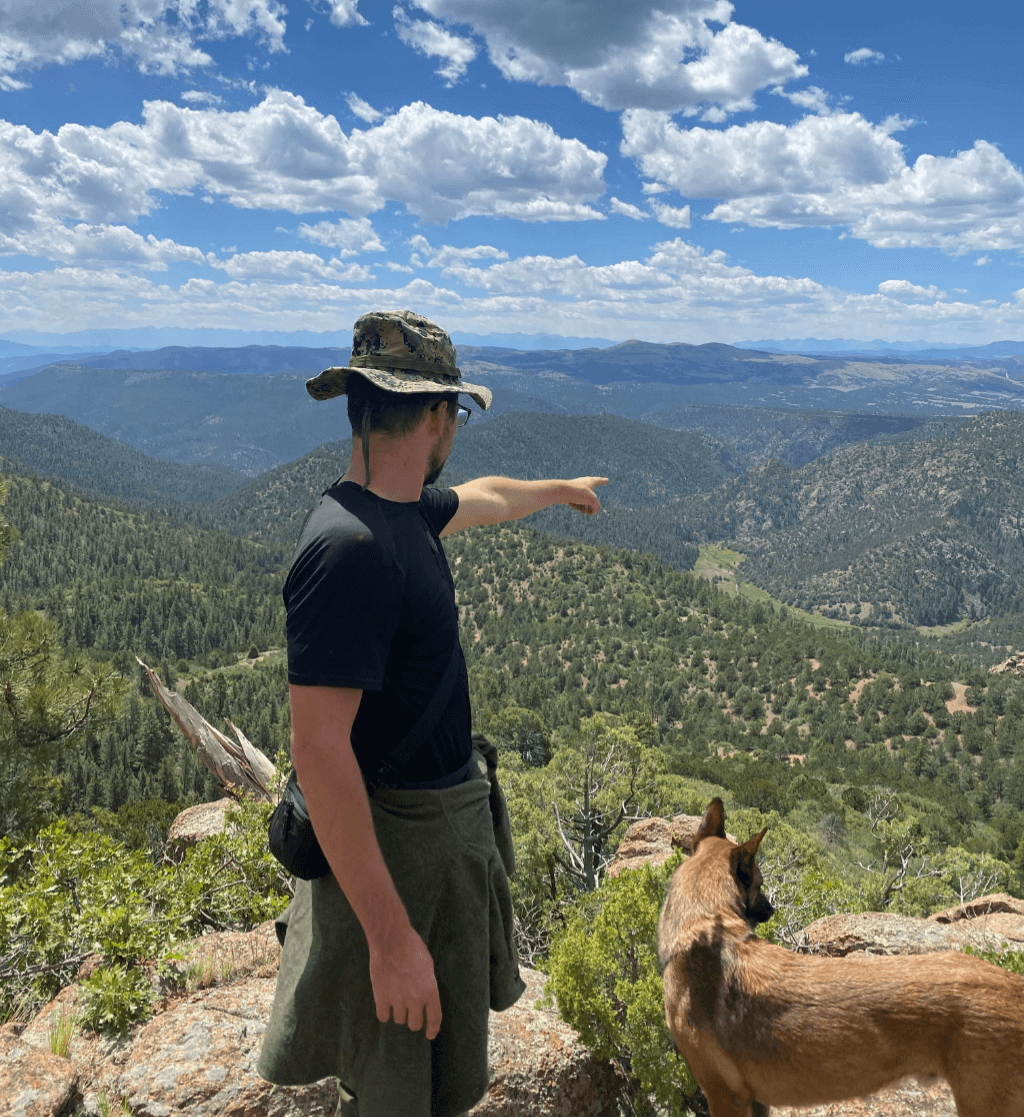 A man wearing a camo boonie hat and black shirt stands on a rocky outcrop pointing into the distance over a vast mountainous forest landscape. Beside him, a Belgian Malinois dog looks out over the canyon. The sky is dotted with fluffy clouds, and layers of green hills stretch out to the horizon under bright daylight.
