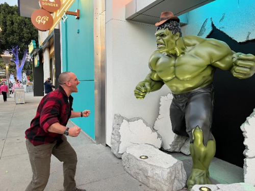 Outdoor scene featuring survival instructor Aaron 'Hutch' Hutchings playfully squaring off against a life-sized Hulk statue emerging from a wall. Hutch's signature Indiana Jones-style hat sits atop the Hulk's head, adding a humorous and personal touch to the encounter.