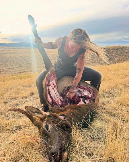 Emily Bartle, instructor at The Survival University, processing an elk in a grassy high plains field at sunset, wearing a black tank top and pants, demonstrating primitive survival and hunting skills training.