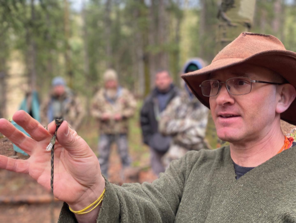 Hutch, instructor at The Survival University, in a brown hat and green coat, holding a cord in a forest, teaching survival skills to a group of students, demonstrating wilderness training expertise.