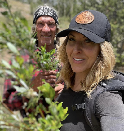 Jess Caldwell, instructor at The Survival University, smiles in a lush Colorado forest with another instructor wearing a bandana and cap, holding green plants, demonstrating wilderness survival and foraging skills training.