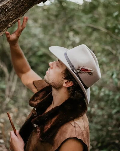 Ryan Maguire, instructor at The Survival University, in a cowboy hat with a feather, touching a tree in a lush forest, wearing a leather vest and holding a stick, showcasing primitive survival skills training.