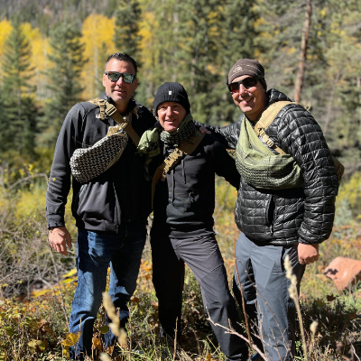Three students, entrepreneurs from across the US, at The Survival University, posing together in a Colorado forest with autumn trees, wearing tactical gear and backpacks, celebrating their epic annual adventure taking a survival course at TSU.