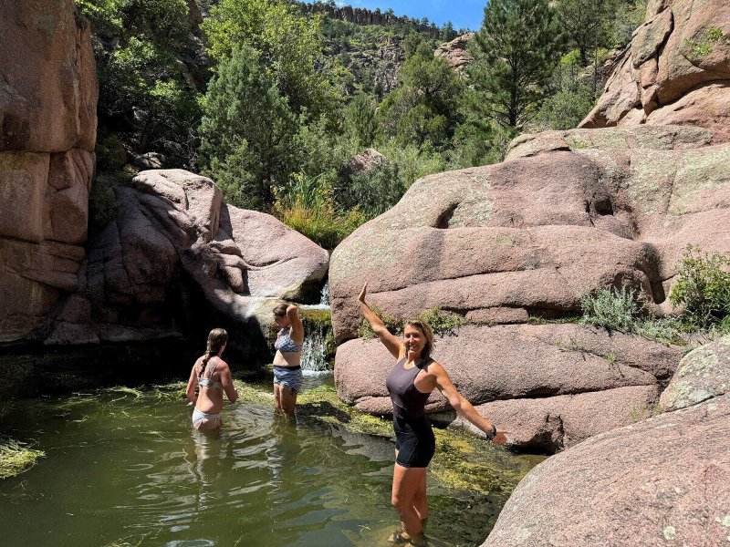 Jess Caldwell, instructor at The Survival University, stands triumphantly in a rocky swimming hole at the Wild Women River Retreat, arms raised, wearing a black swimsuit. Two students wade nearby in the clear water, surrounded by Colorado’s rocky canyons and lush greenery, showcasing TSU’s wilderness immersion training.