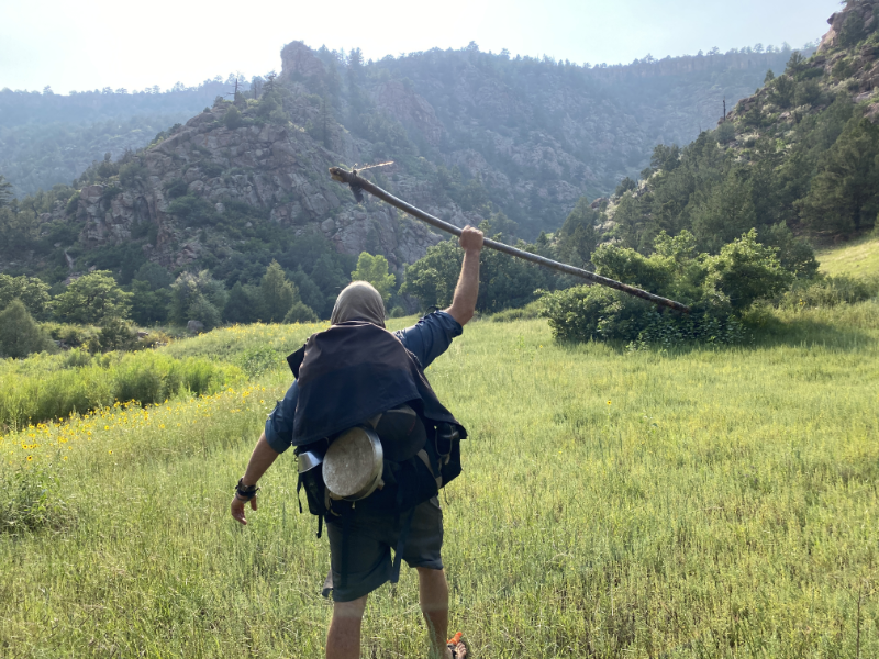 Student at The Survival University, celebrating in a grassy Colorado valley with jagged mountains in the background, holding a long stick, wearing a headscarf, dark jacket, and backpack, after a challenging hike nearly reaching the day’s destination.
