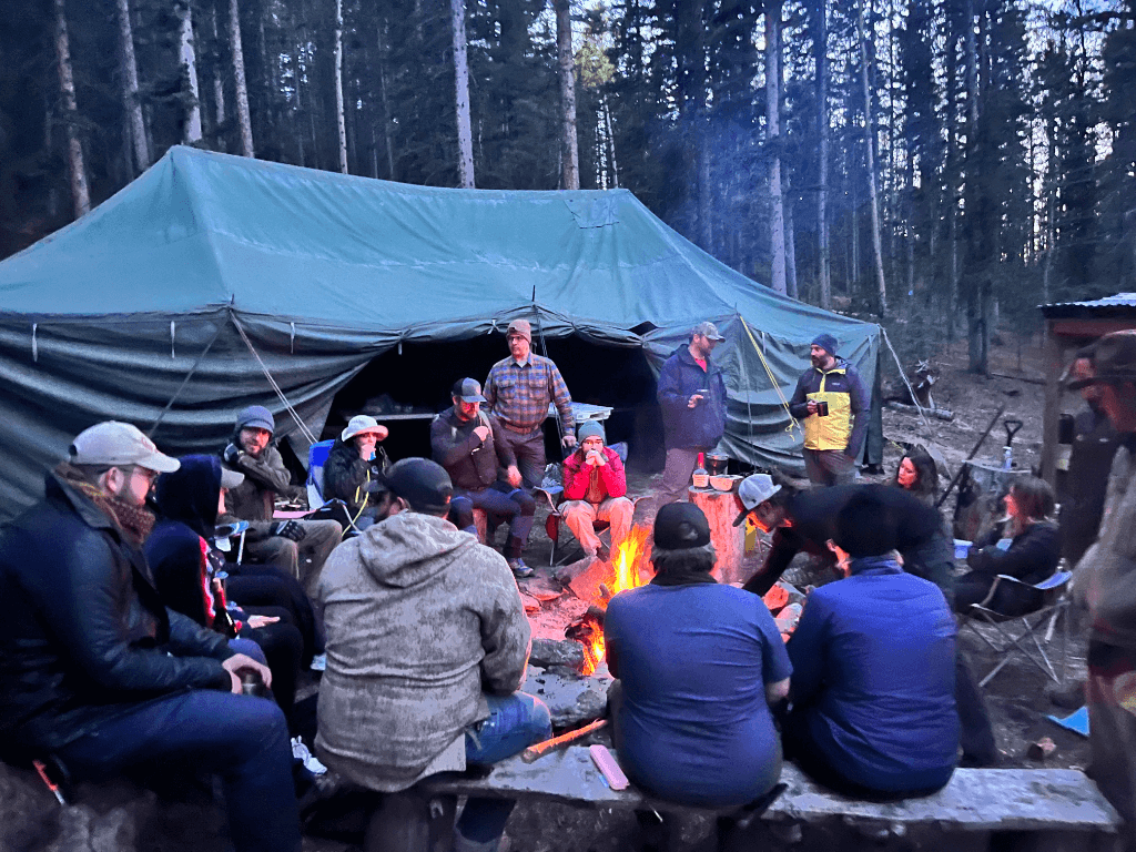 A group of people gather around a glowing campfire outside a large green canvas tent, deep in a pine forest at dusk. The firelight illuminates their faces as they sit on benches and camp chairs, sharing stories, laughter, and warm drinks. Some stand chatting while others lean in close to the fire, wrapped in outdoor layers, forming a circle of connection and camaraderie in the wilderness.