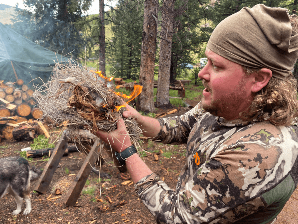 A man in camouflage clothing and a tan headwrap holds a burning tinder bundle made of dry grass and bark, demonstrating fire-starting techniques in a forest setting. Behind him, a green canvas tent and a stacked woodpile suggest a rustic wilderness camp. A dog wanders nearby as the scene captures a hands-on moment of primitive firecraft during outdoor training.