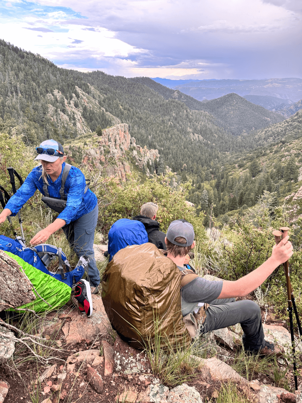 A group of hikers take a break on a rocky mountainside trail, surrounded by dense forest and dramatic cliffs in the Colorado wilderness. One woman in a blue shirt adjusts gear near colorful backpacks, while others sit resting with trekking poles and rain covers over their packs. The sky is overcast, hinting at an approaching storm, and the expansive view stretches across layers of rugged, pine-covered peaks.